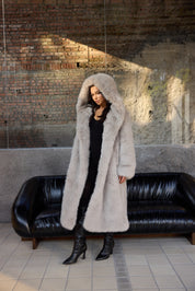 Person wearing a long fur coat standing in front of a black leather couch with a brick wall background.