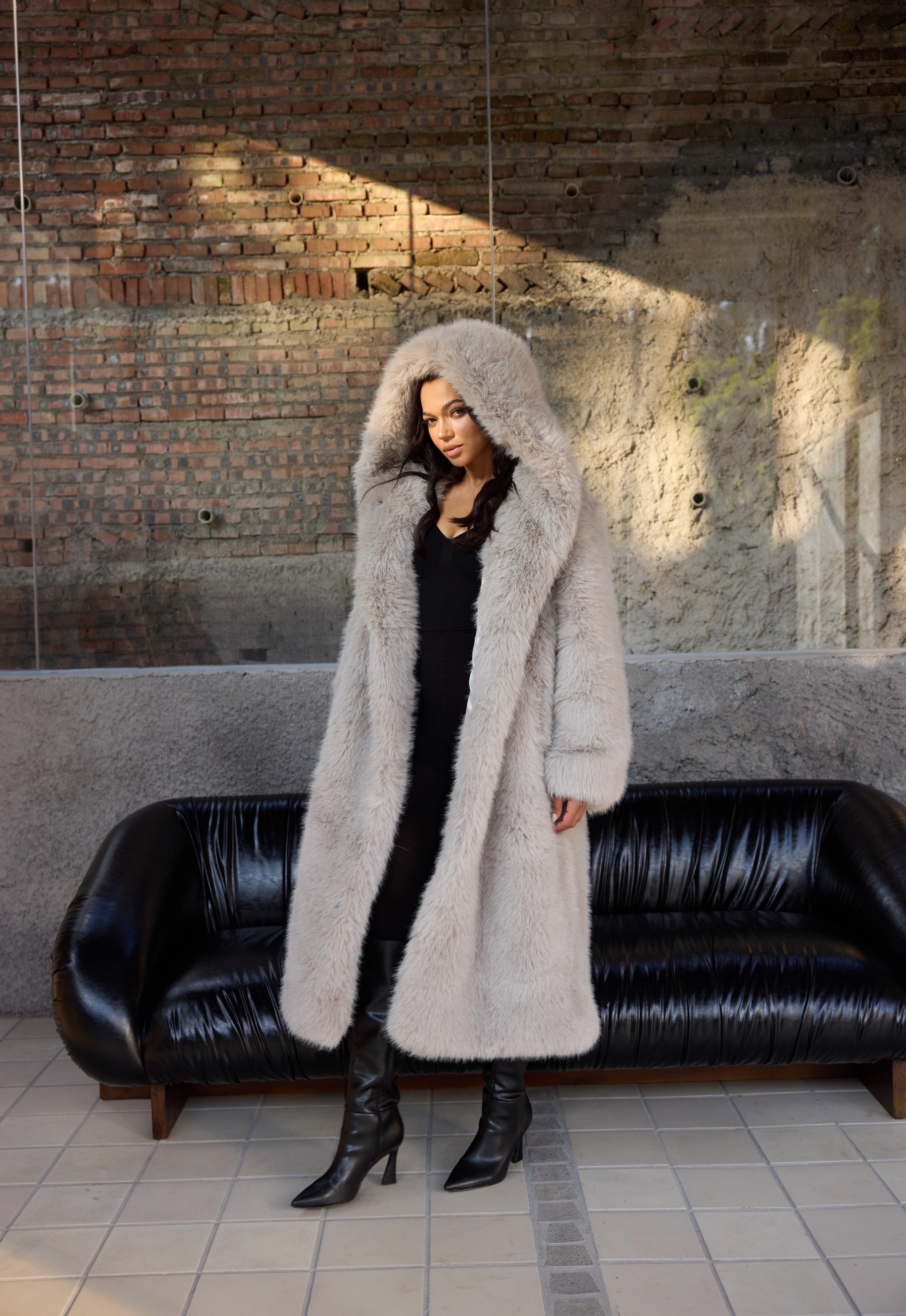 Person wearing a long fur coat standing in front of a black leather couch with a brick wall background.