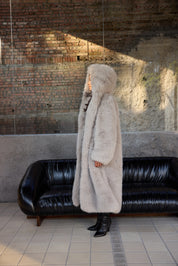 Person wearing a long fur coat standing in front of a black leather couch against a brick wall.