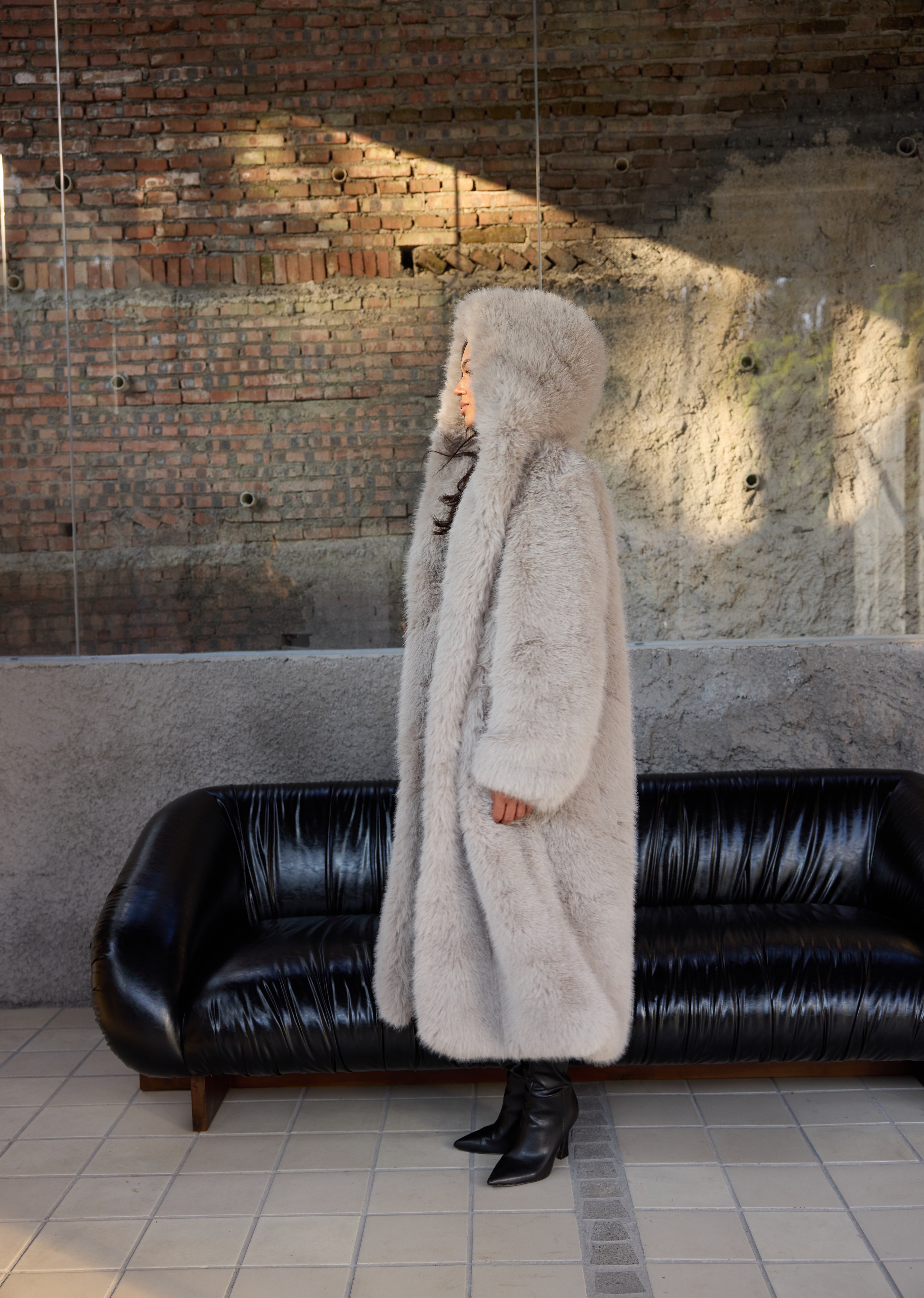 Person wearing a long fur coat standing in front of a black leather couch against a brick wall.