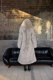 Person wearing a long fur coat standing in front of a black sofa against a brick wall.
