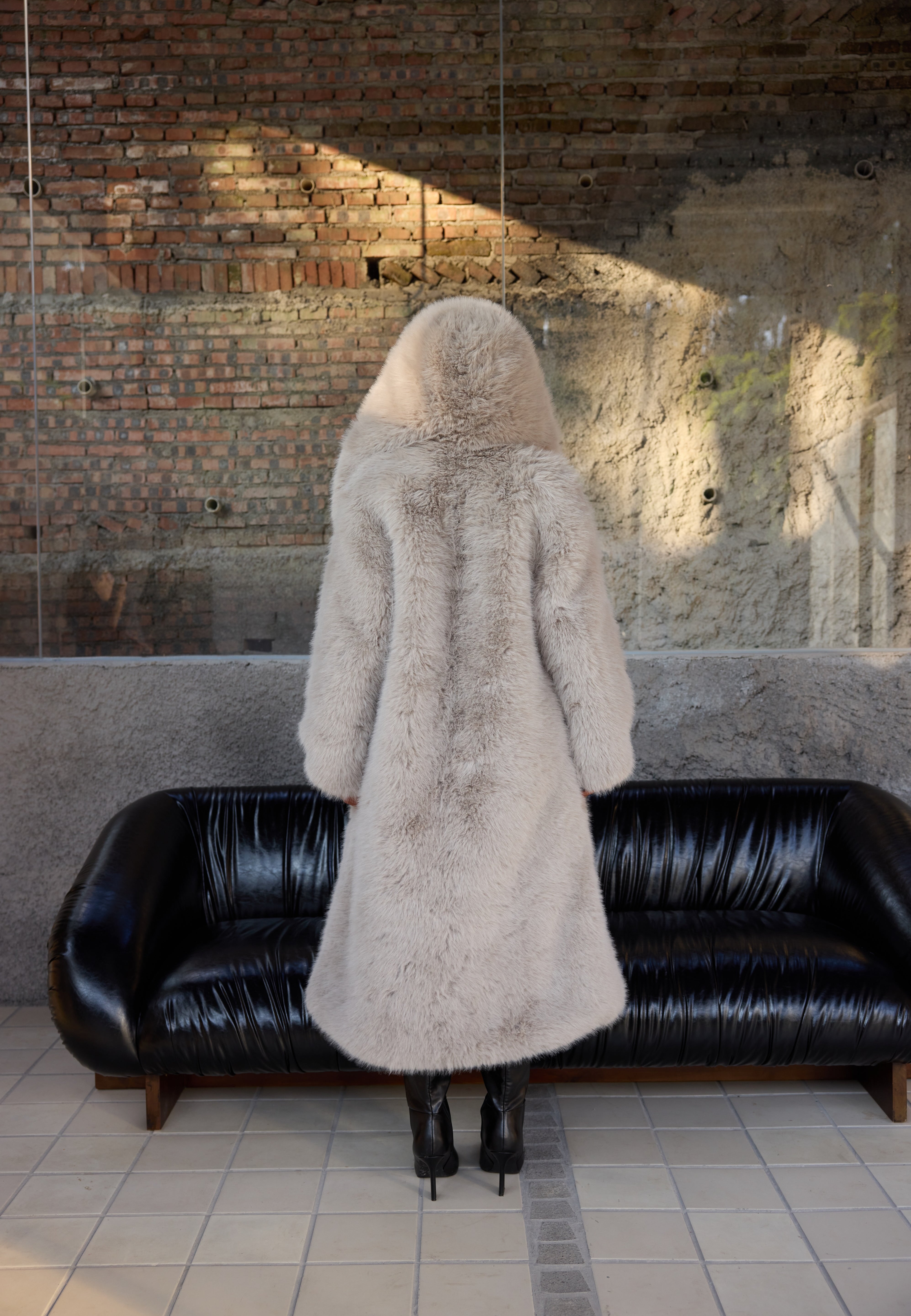 Person wearing a long fur coat standing in front of a black sofa against a brick wall.
