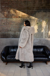 Woman in a long fur coat standing in a room with a black leather couch and brick wall.