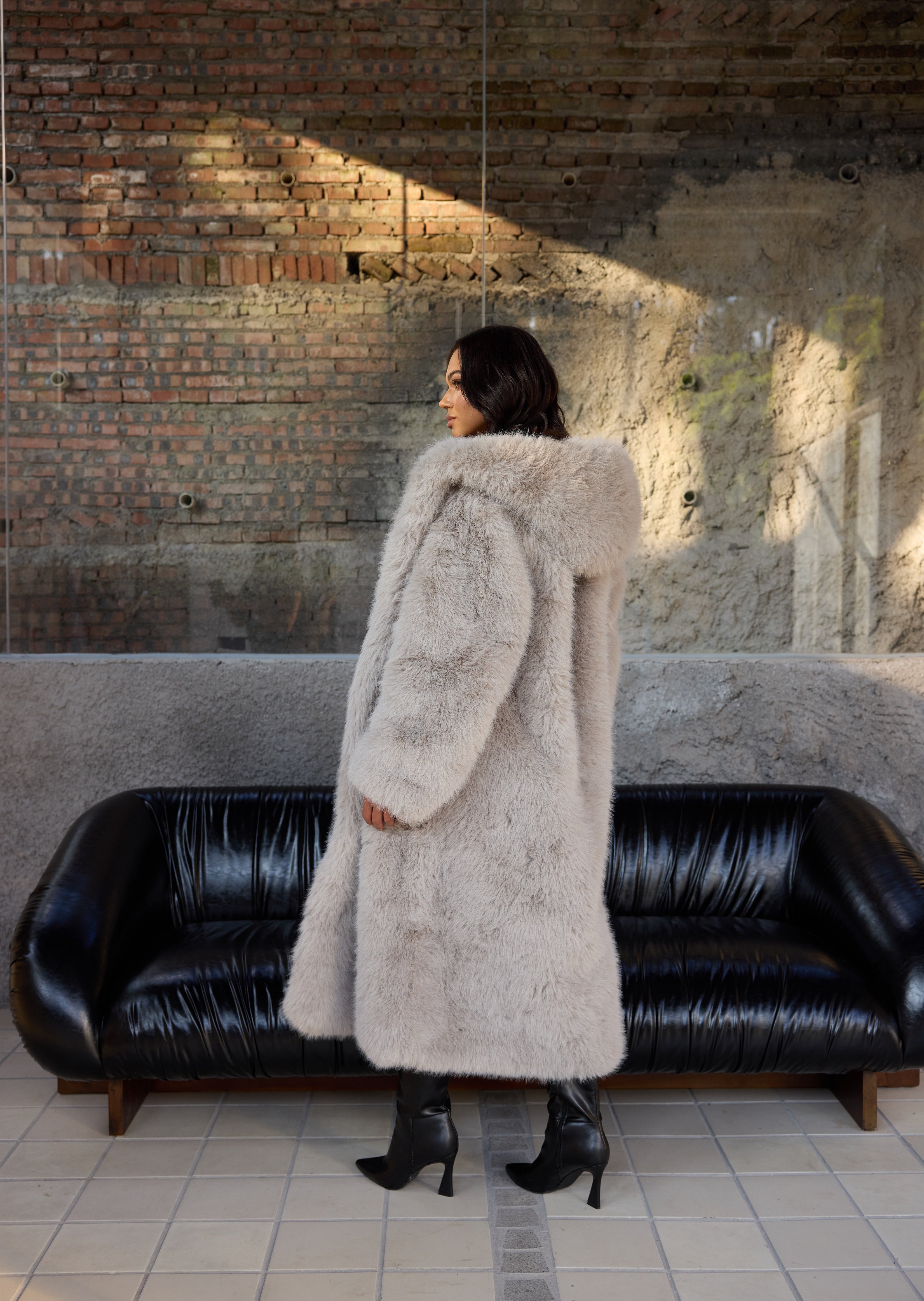 Woman in a long fur coat standing in a room with a black leather couch and brick wall.