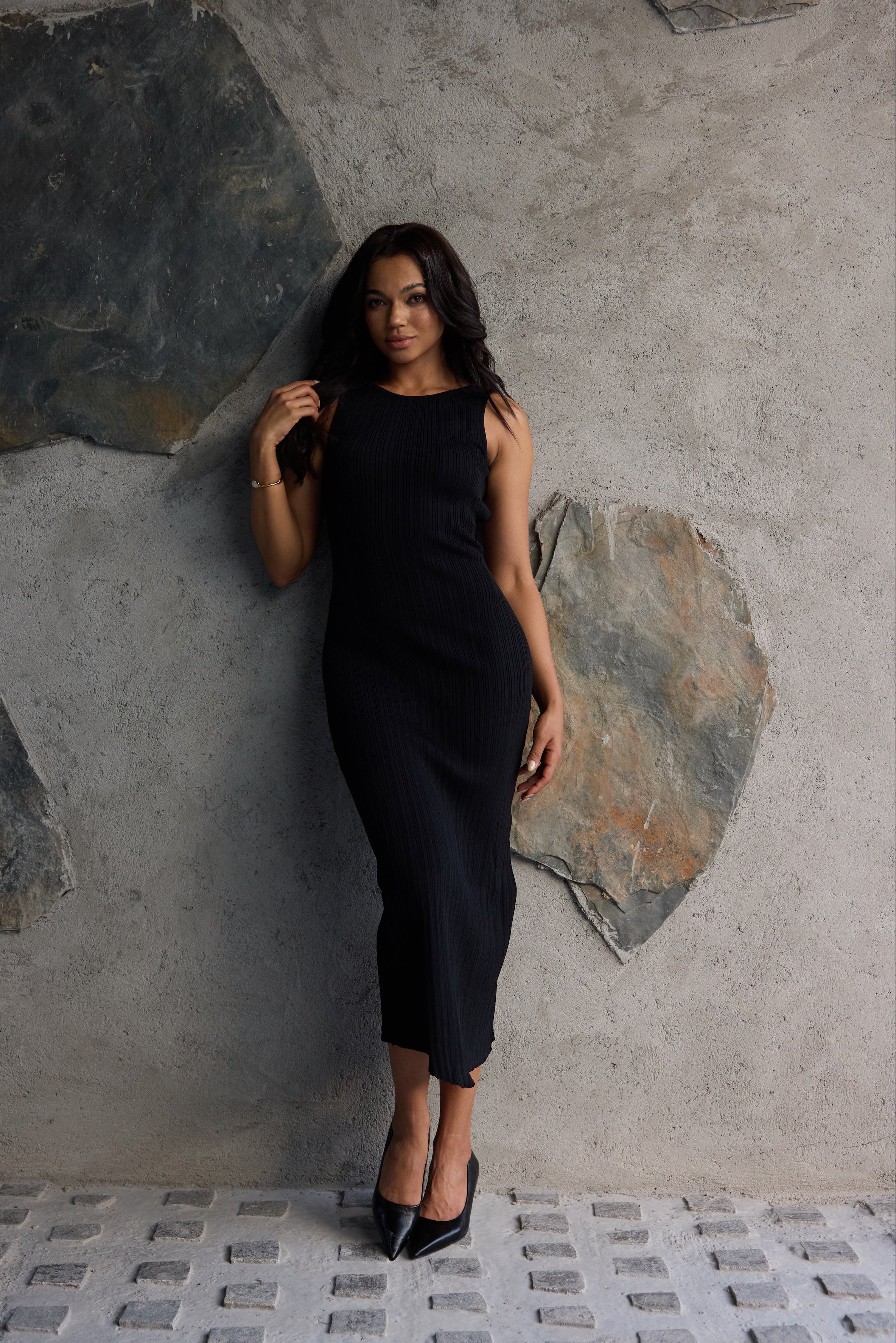 Woman in a black dress standing against a textured stone wall.