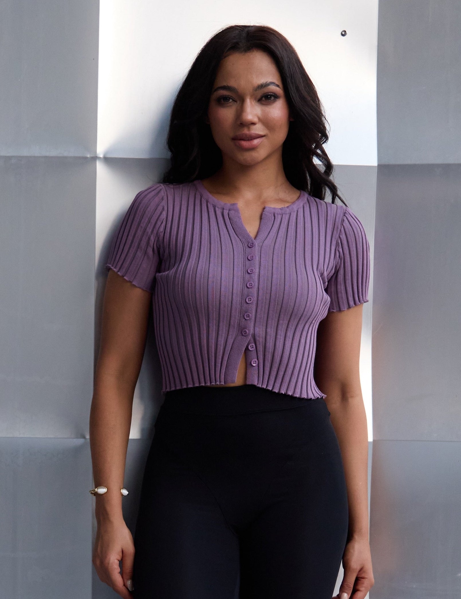 Woman in a purple top and black pants standing against a geometric wall.