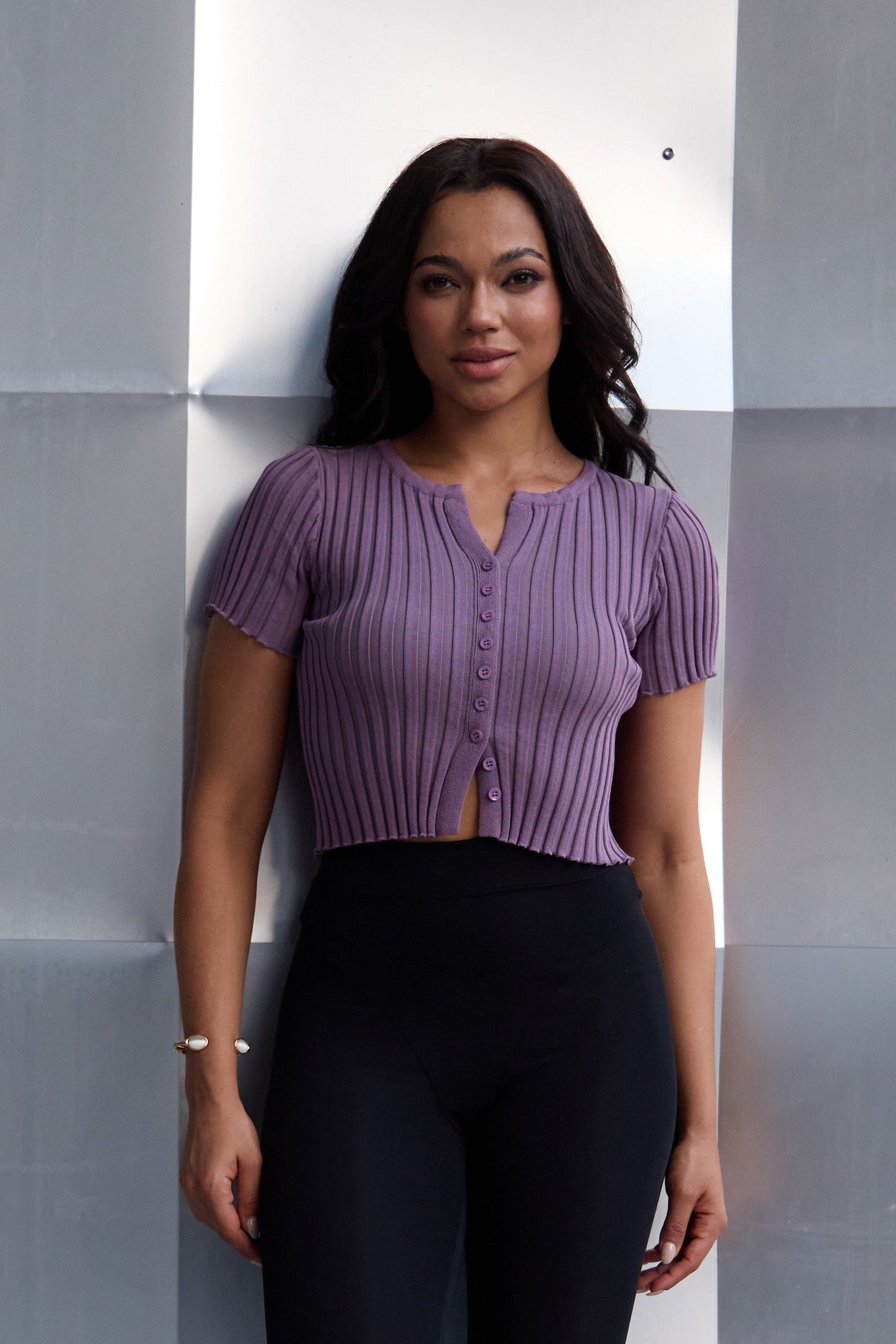 Woman in a purple top and black pants standing against a geometric wall.