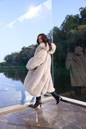Woman in a fur coat standing by a reflective surface with trees in the background