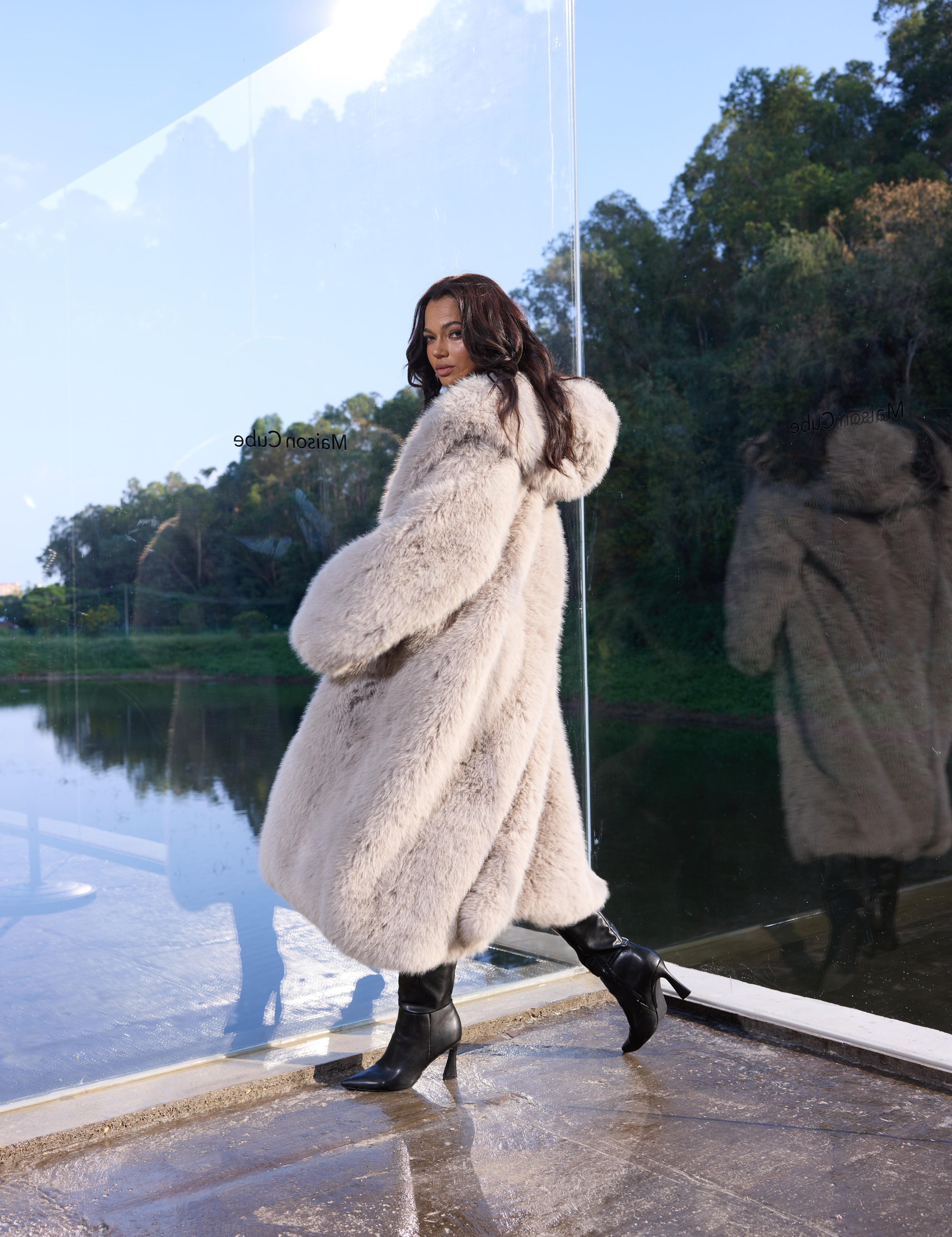 Woman in a fur coat standing by a reflective surface with trees in the background
