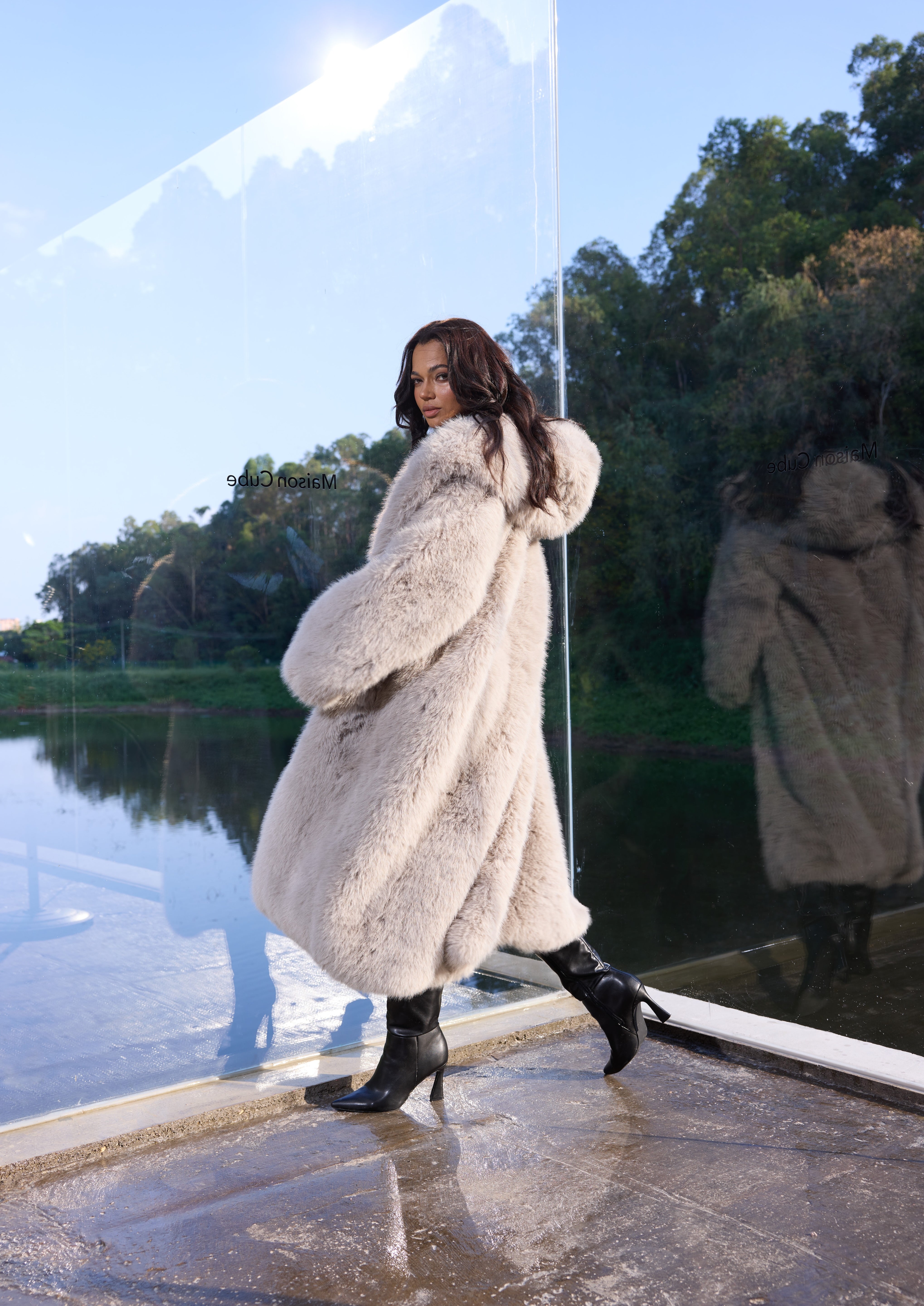 Woman in a fur coat standing by a reflective surface with trees in the background